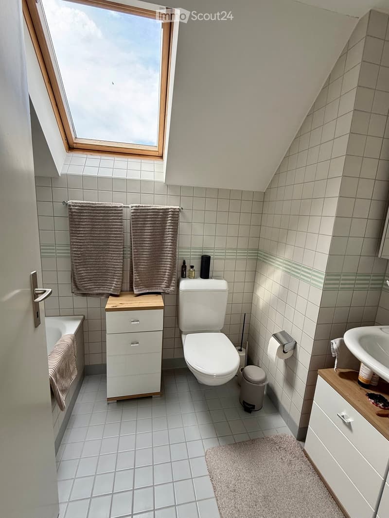 Bright, compact bathroom with white tiles, a toilet, sink, bathtub, and wooden cabinet. Features two towels, a trash bin, and a small rug on the floor.