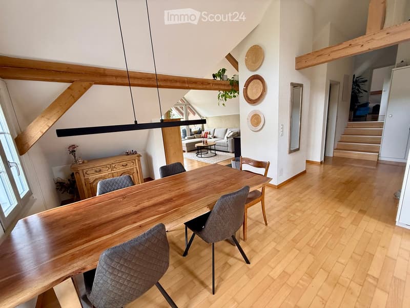 Open-plan dining and living area with wooden floors, a long wooden dining table with four chairs, a grey sofa, a coffee table, and a modern pendant light fixture.