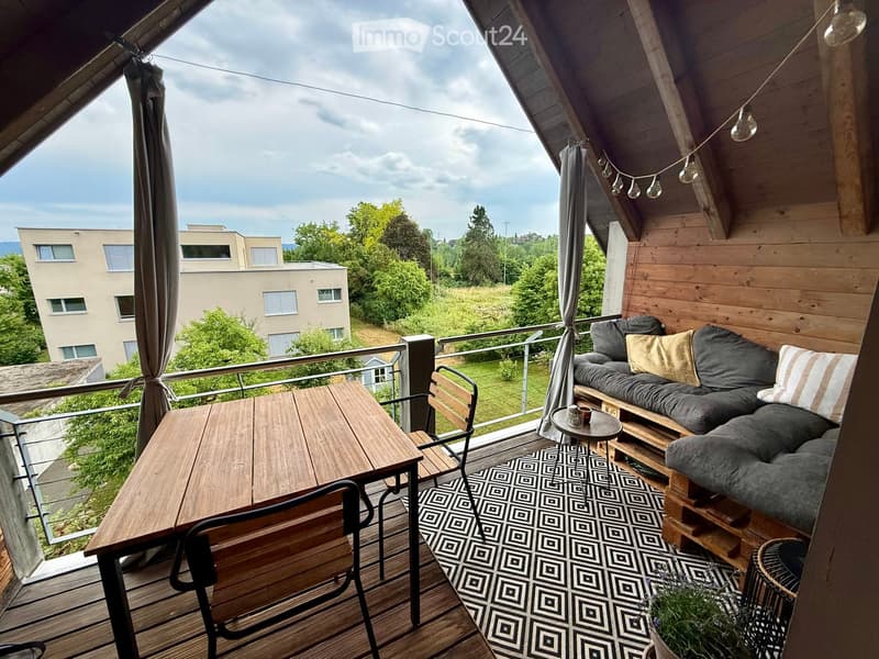 Wooden balcony with a grey sofa, small round table, wooden chairs, patterned rug, and string lights.