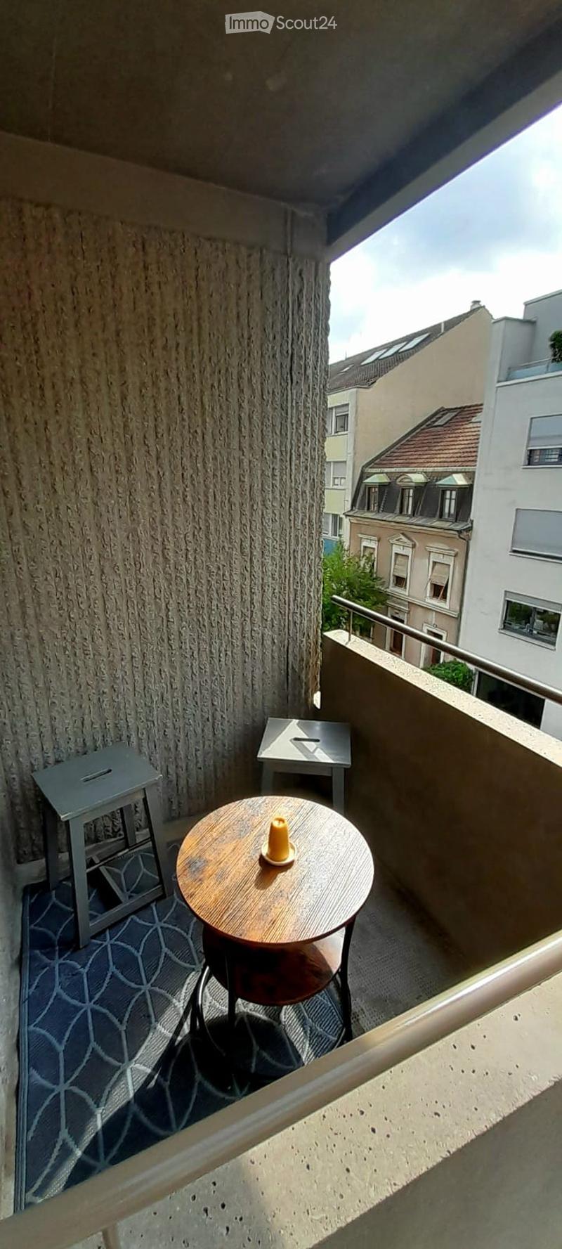Balcony with a round wooden table, two stools, and a small decorative item on the table. The balcony has a textured wall and a patterned rug on the floor.