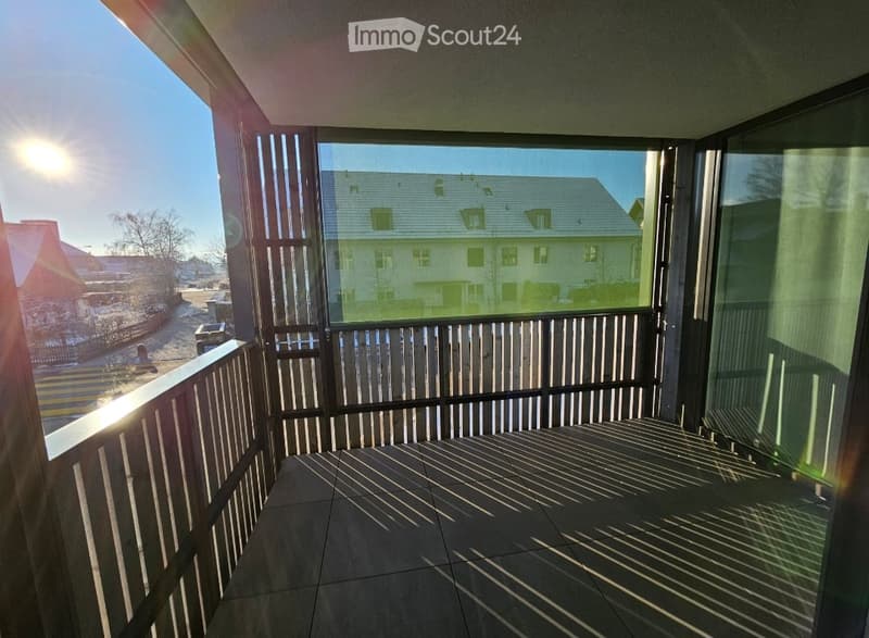 Balcony with tiled floor, wooden railing, large windows offering a view of a residential area with houses and trees.