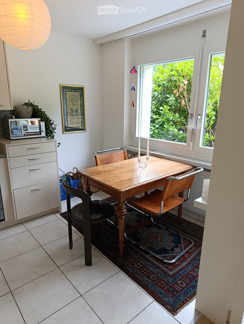 Bright room with a wooden dining table, chairs, microwave, cabinet, and a framed picture on the wall. Natural light from large windows and a view of greenery outside.