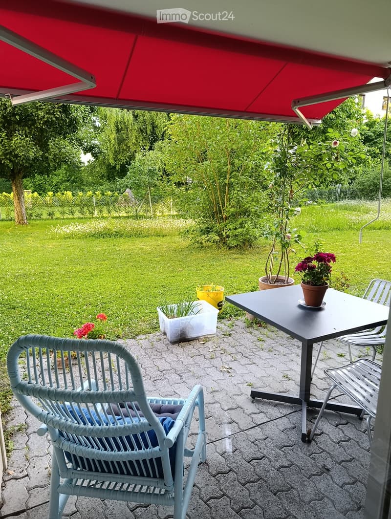 Outdoor patio with a table, chairs, and potted plants, surrounded by a well-maintained garden with grass and trees.