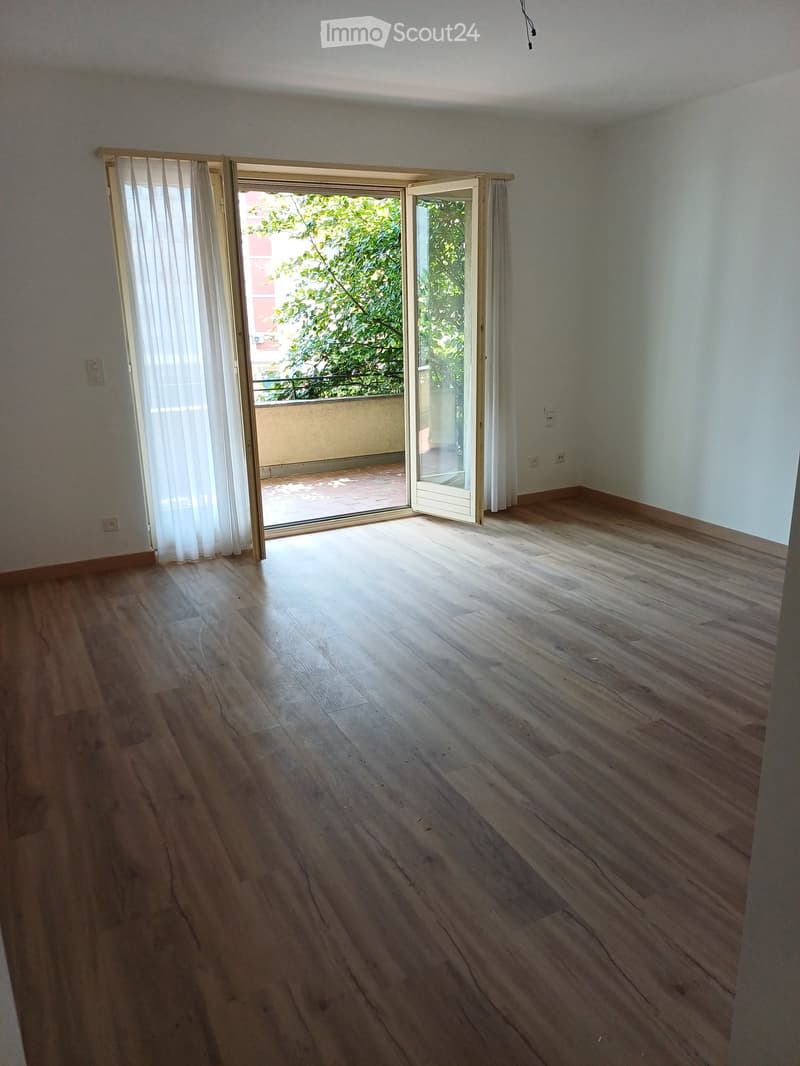 Empty room with wooden flooring, white walls, and large sliding glass doors leading to a balcony with outdoor space and greenery visible.