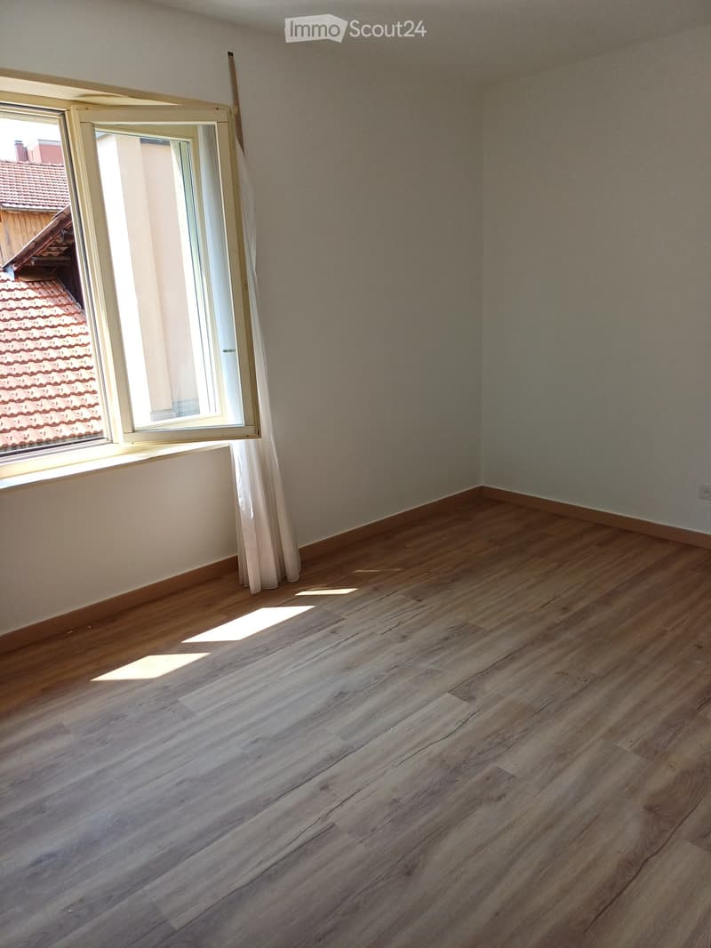 Empty room with wooden flooring and large window showing a neighboring roof. Bare walls with no furniture or decorations.
