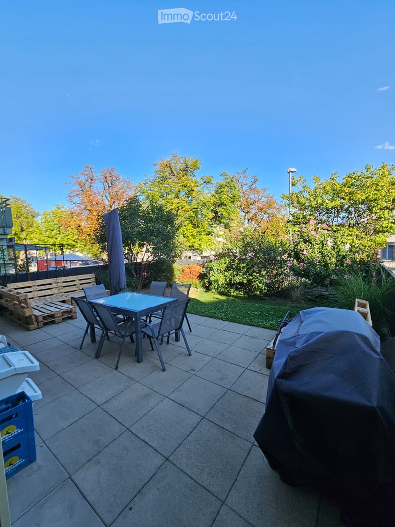 Outdoor patio with tiled floor, a table and chairs, a covered grill, a bench, and surrounding greenery.