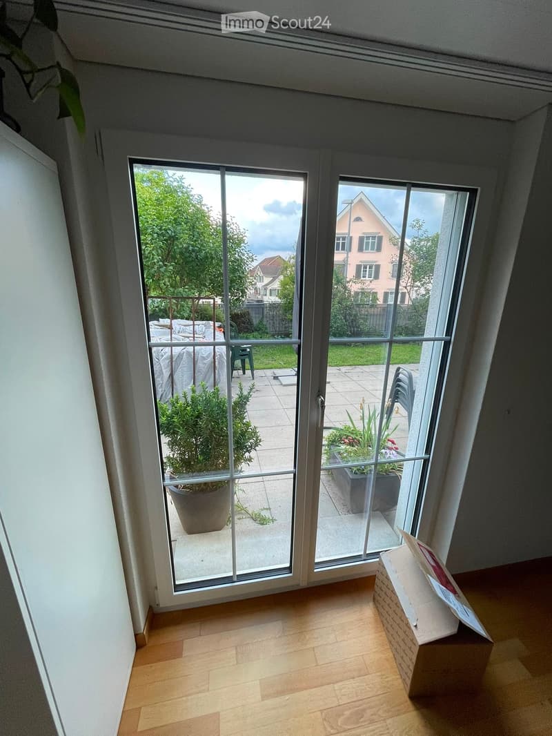 Empty room with wooden flooring, large glass sliding doors leading to a patio with potted plants and outdoor furniture.