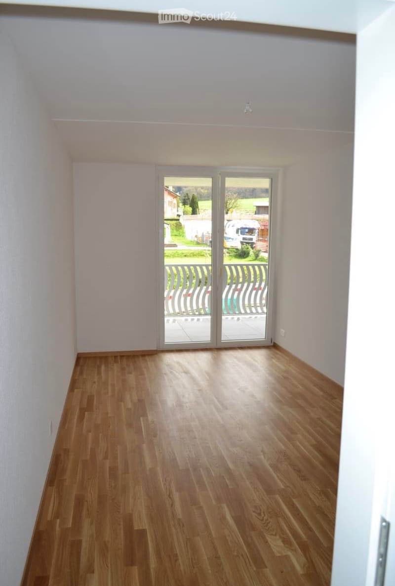 Empty room with light wooden flooring and white walls, featuring a large sliding glass door leading to a balcony with a view of the outdoors.