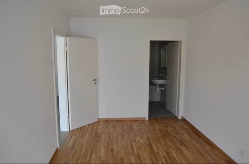 Empty room with light-colored walls and wooden flooring, featuring an open door leading to a bathroom with a sink and mirror.