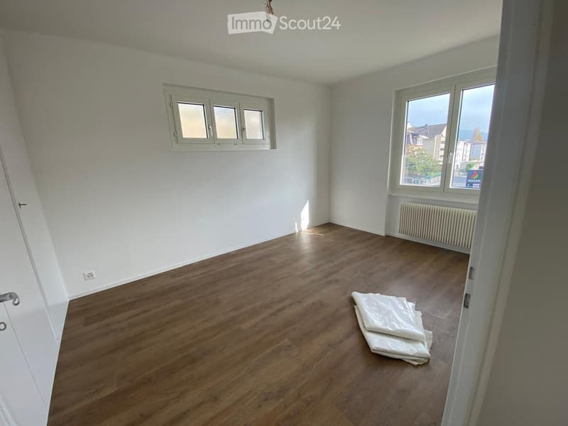 Empty room with wooden flooring, white walls, and two windows. A radiator is visible under one window, and a folded white item is on the floor.