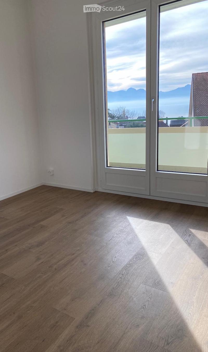 Empty room with wooden flooring and large windows offering a scenic view of mountains and a lake.