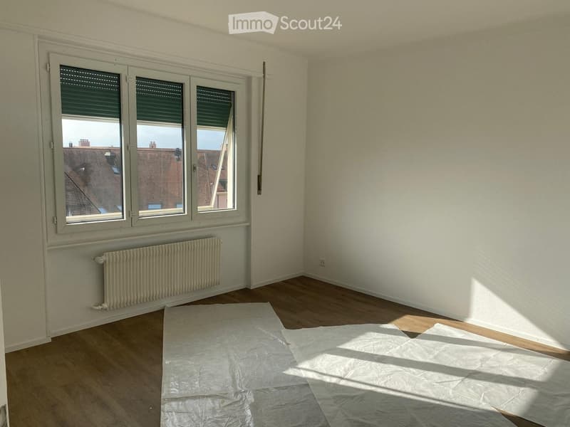 Empty room with wooden flooring, white walls, large windows with blinds, radiator, and protective plastic sheets on the floor.