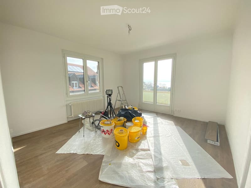 Empty room with wooden flooring, white walls, large windows, and a radiator. The floor is covered with protective plastic sheeting, and various construction materials and tools are scattered around.