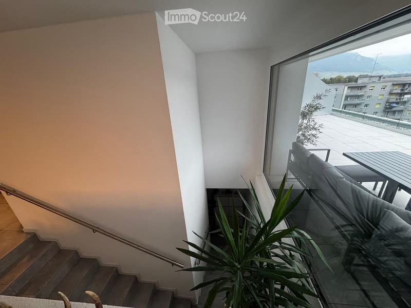Modern hallway with a staircase leading upwards, large window offering a scenic mountain view, and a potted plant near the window.