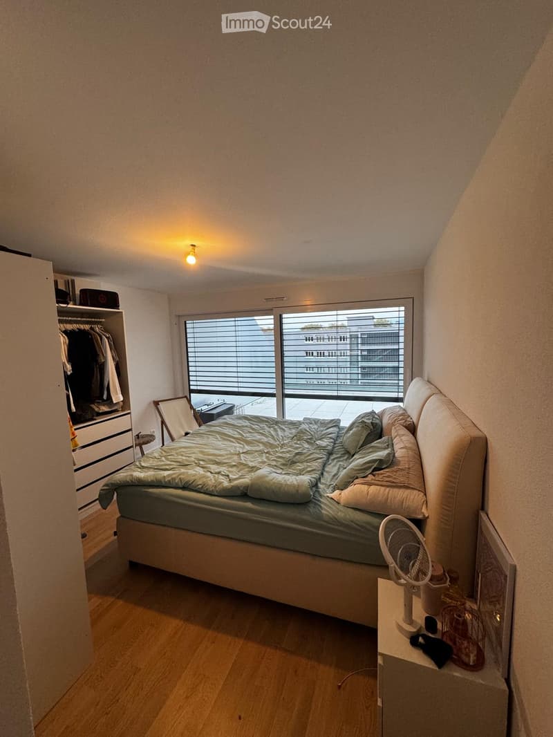 Bedroom with a large bed, nightstand, fan, wardrobe, and a mirror. The room has wooden flooring and large windows with blinds.