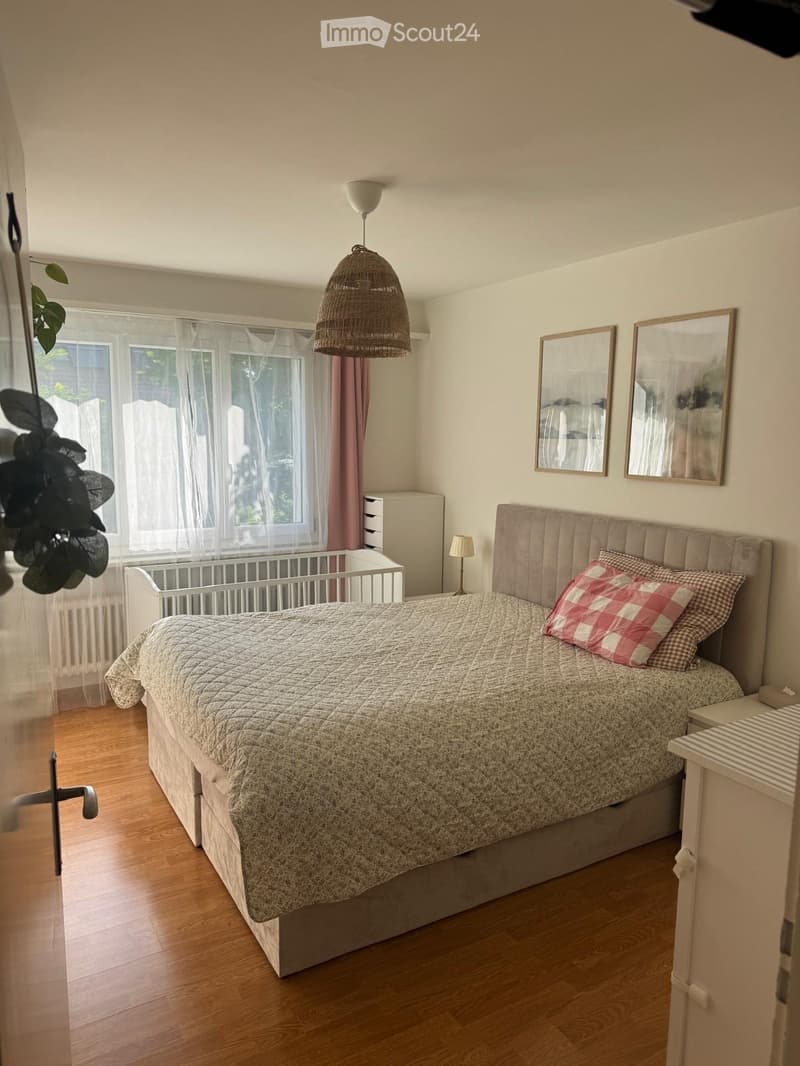 Cozy bedroom with wooden flooring, large window with white curtains, a double bed with a patterned quilt and pillows, a nightstand with a lamp, two wall-mounted mirrors, a wicker pendant light, a small plant, and a baby crib near the window.