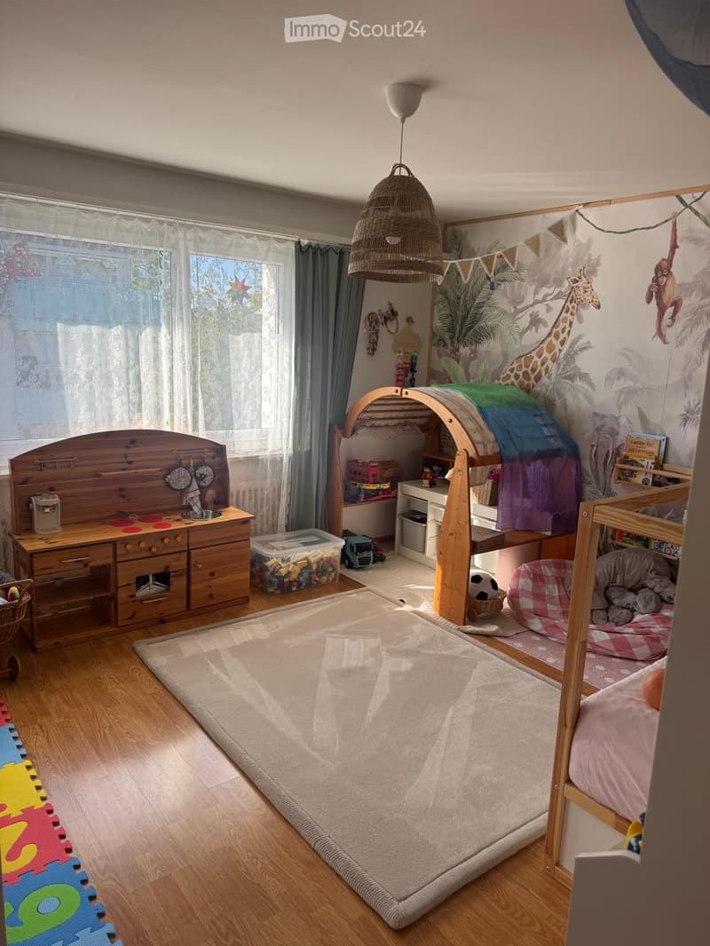 A well-lit children's bedroom with a bunk bed, a toy kitchen, a hanging wicker lamp, and a large window with curtains. The room features a colorful play mat and a variety of toys.