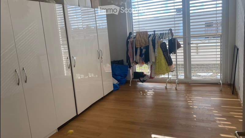 Empty room with wooden flooring, white built-in wardrobes, and a clothes drying rack near the window. The room is well-lit with natural light coming through large windows with blinds.