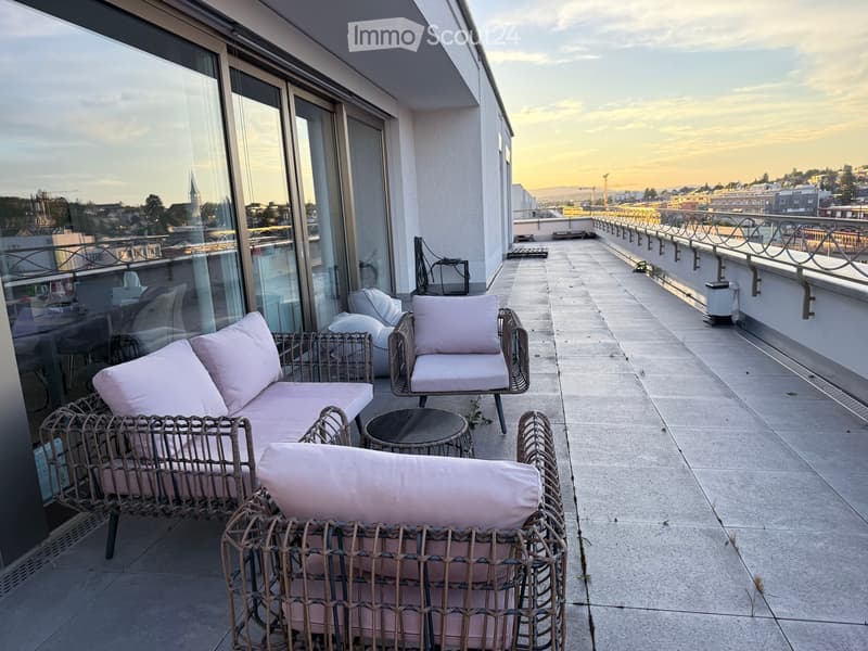 Balcony with wicker furniture, including a sofa, chairs, and a small table, overlooking a cityscape at sunset.