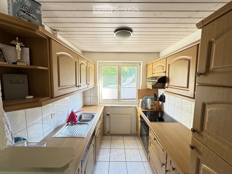 The kitchen features light wooden cabinets, a central island, and a sink with a faucet. Appliances include an oven and stove, and there are various kitchen items such as a cup, knife block, and a radiator under the window.