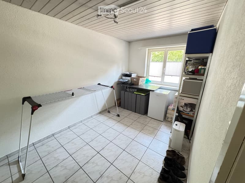 A well-lit utility room with tiled flooring, a drying rack, a refrigerator, a washing machine, a table with a chair, storage boxes, a ladder, and a shoe rack.