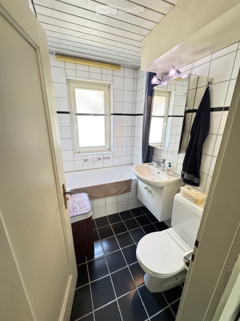 Bathroom with white tiles, black floor tiles, bathtub, sink, toilet, mirror, and a towel hanging on the wall.