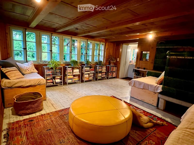 Cozy living room with wooden walls and ceiling, large windows, a fireplace, a round ottoman, a sofa, bookshelves, and a woven basket.