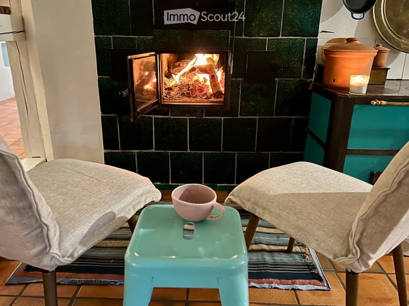 Cozy living space with a lit fireplace, two chairs, a small green stool holding a pink cup, and a candle on a side table with a pot.