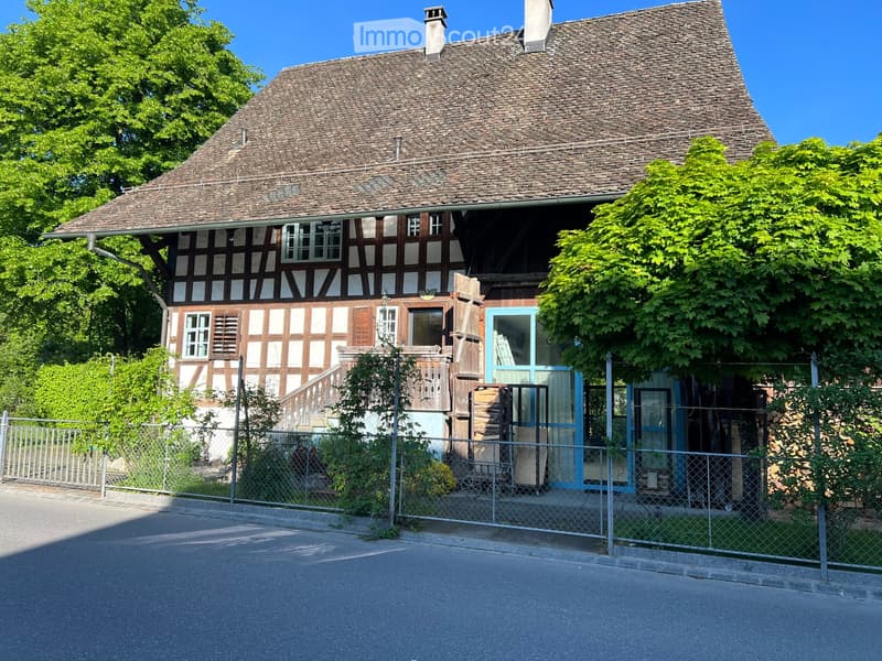 Two-story traditional half-timbered house with a steeply pitched roof, white and brown facade, multiple windows, and a small porch with a staircase leading to the entrance.