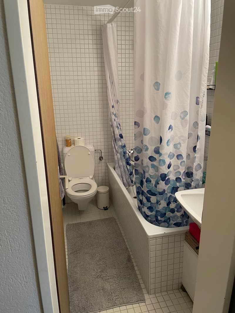 Compact bathroom with a toilet, bathtub with a shower curtain, sink, and a small trash can. The walls are tiled, and a bath mat is placed on the floor.