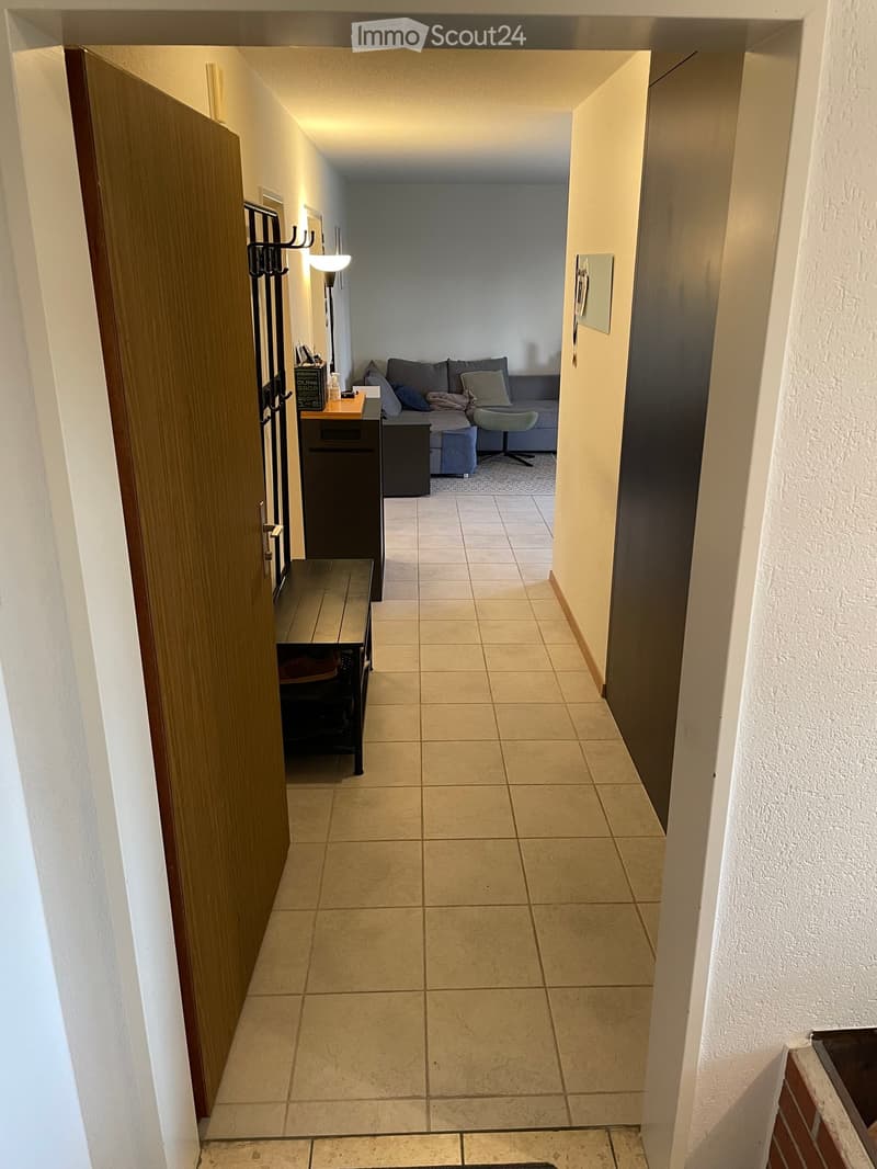 Hallway with tiled floor, wooden door on the left, and a view into a living room with a sofa and small table.