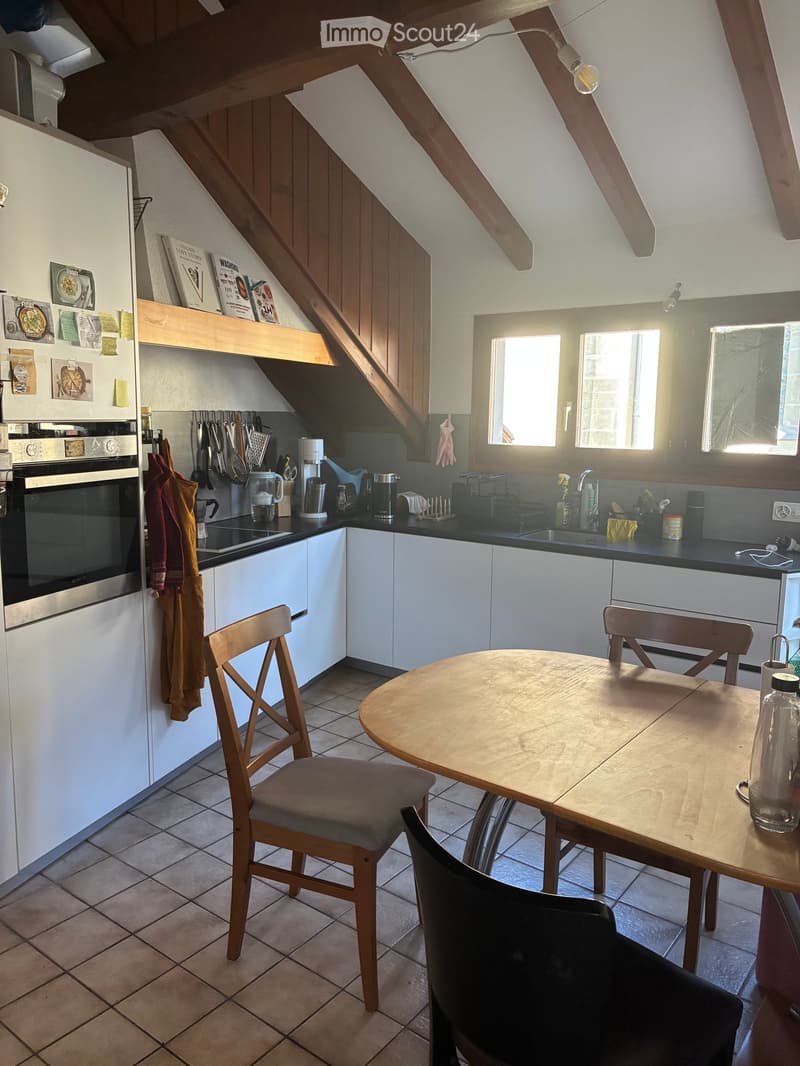Bright kitchen with white cabinets, stainless steel appliances, and a wooden dining table with chairs. The space features a tiled floor and large windows allowing natural light to fill the room.