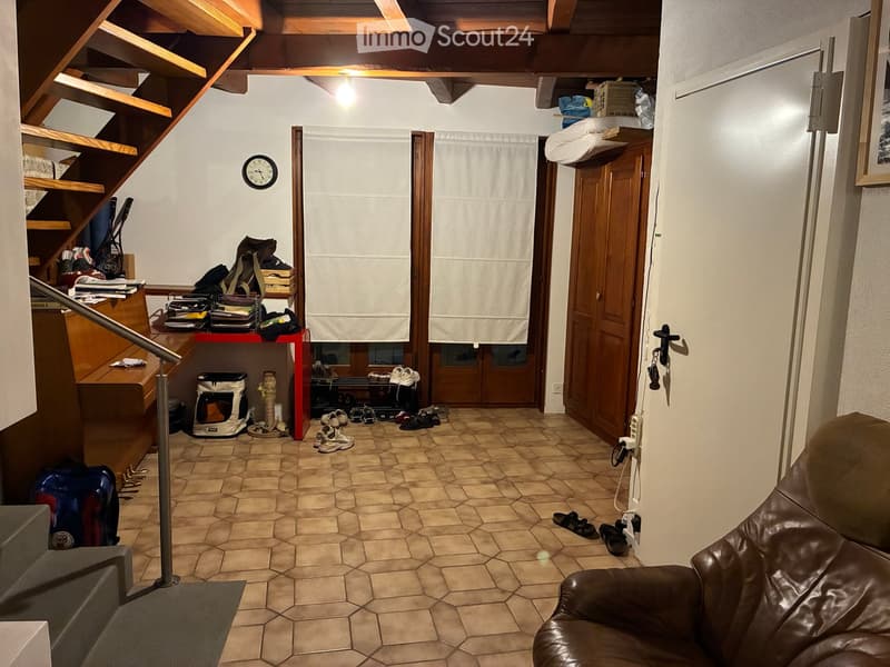 A cozy hallway with a wooden staircase, a red table with shoes and bags, a pet carrier, and a brown leather sofa on the right side.