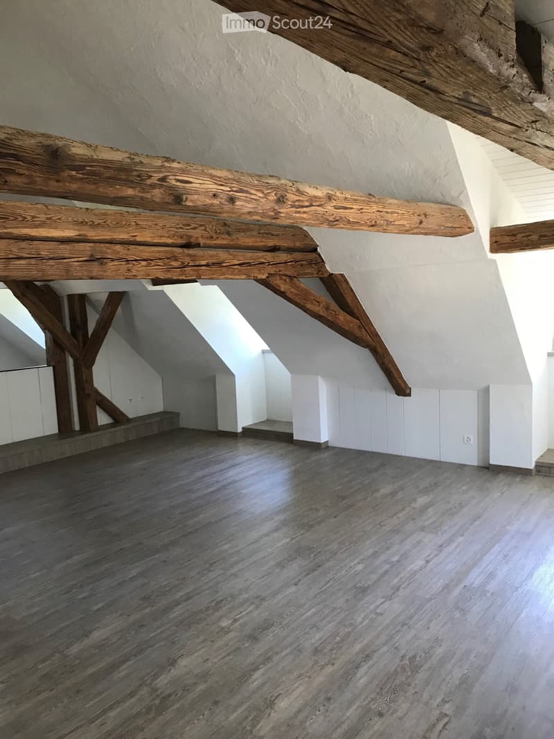 Empty attic space with exposed wooden beams, wooden flooring, and white walls. No furniture or equipment present.