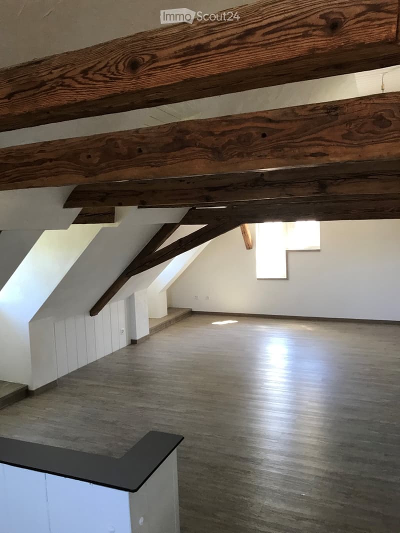 Empty attic space with exposed wooden beams, white walls, and a wooden floor. Natural light streams in from a skylight window.