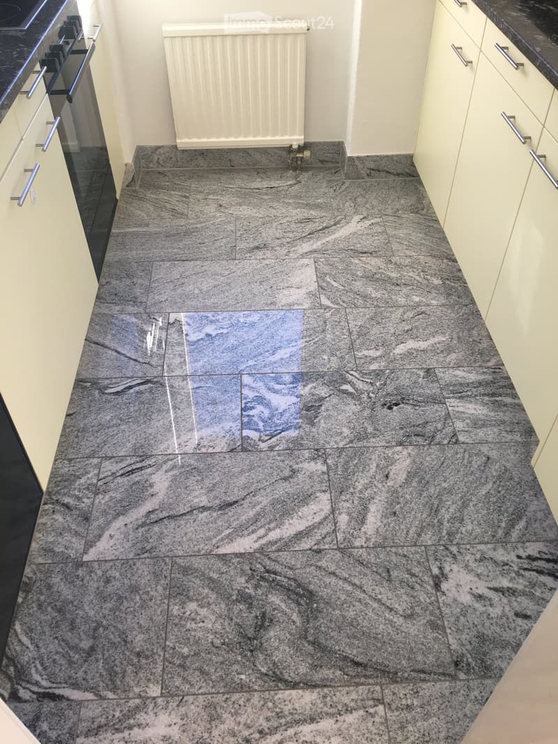 Close-up view of a kitchen floor with large, polished grey marble tiles arranged in a herringbone pattern. The tiles have a polished finish and a subtle veining pattern. The surrounding area includes cream-colored cabinetry with silver handles and a radia