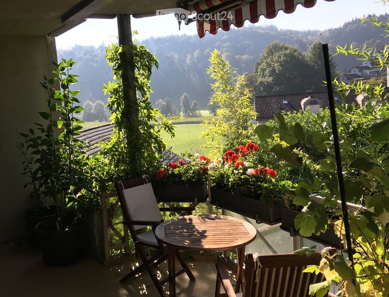 Balcony with a round wooden table, two chairs, and potted plants, overlooking a scenic valley with houses and greenery.