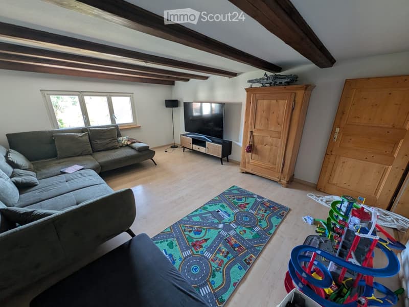 Living room with exposed wooden beams, grey sectional sofa, television on stand, floor lamp, toy car track rug, and various toys.