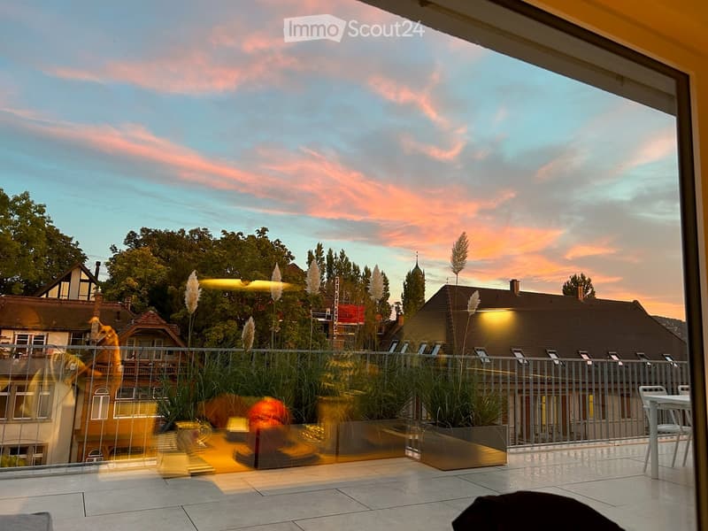 Balcony with modern glass railing, tiled floor, potted plants, and outdoor furniture, overlooking a residential area with houses and trees during sunset.