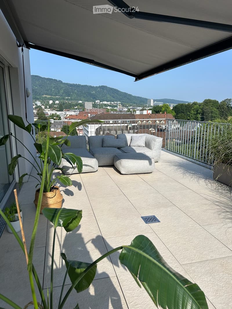 Balcony with modern furniture, including a grey sectional sofa and ottoman, surrounded by potted plants, offering a scenic city and mountain view.