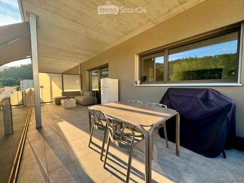 Balcony with modern furniture including a sofa, small table, dining table with chairs, and a covered grill. Large windows offer a scenic view of greenery.