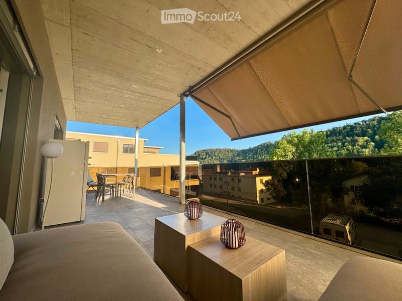 Modern balcony with wooden ceiling, large glass railing, and scenic view of greenery and buildings. Features a sofa, small table with decorative lamps, and outdoor dining set.