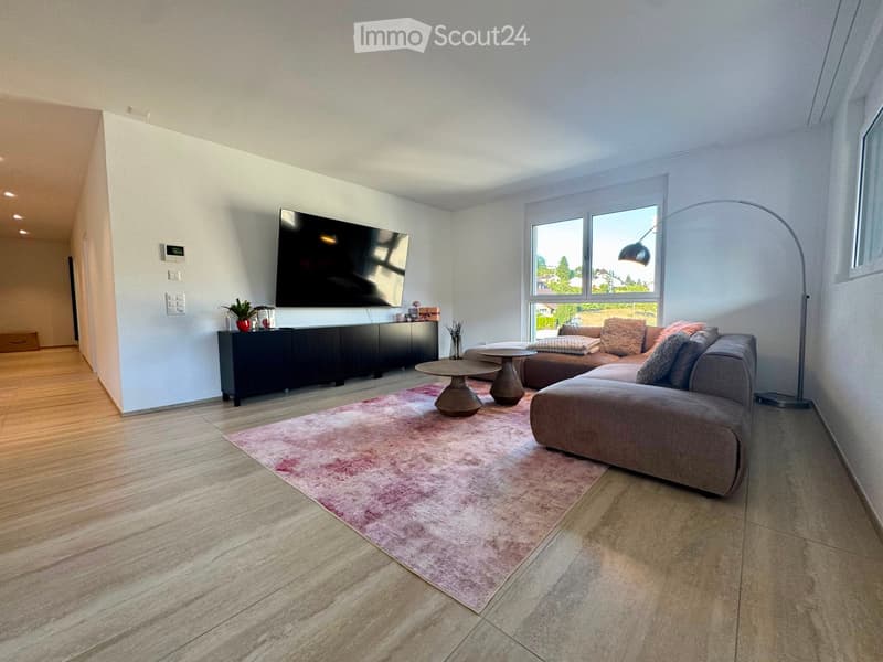 Modern living room with a grey sectional sofa, coffee table, TV mounted on the wall, floor lamp, and a pink area rug on a tiled floor.