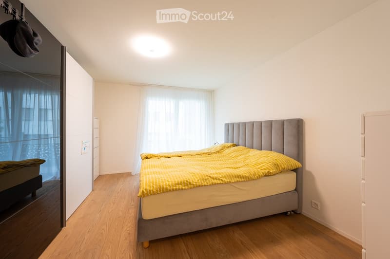 Bed with a grey headboard, yellow bedding, a ceiling lamp, and white walls. There is a window with curtains. The room also features a mirrored wardrobe and wooden floor.