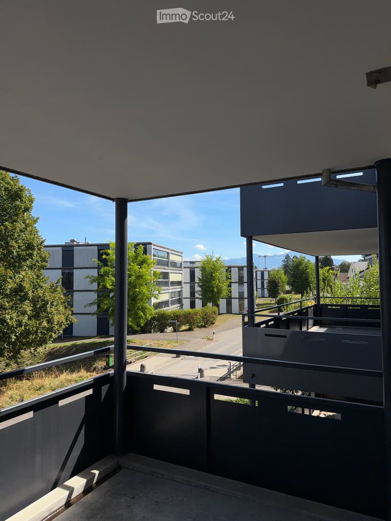 Balcony with metal railing, overlooking a modern residential complex with trees and a clear blue sky.