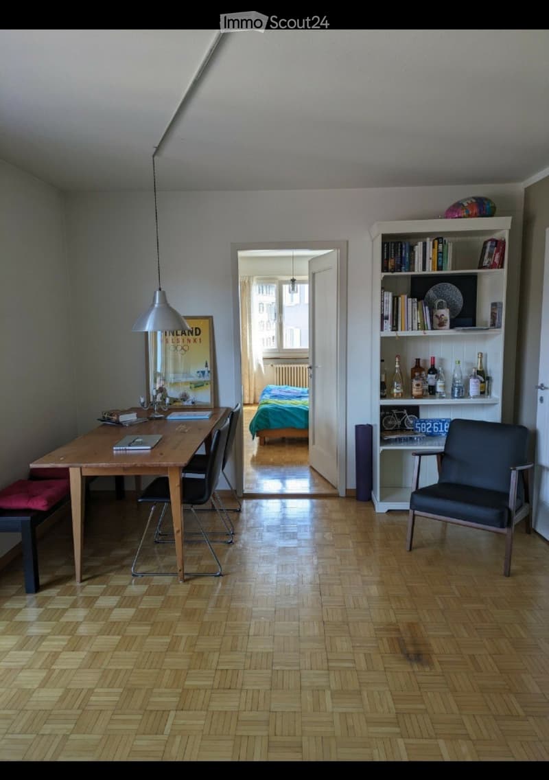 A well-lit room with wooden flooring, a dining table with chairs, a bookcase, a mirror, and a lamp. An adjacent bedroom is visible through an open doorway.