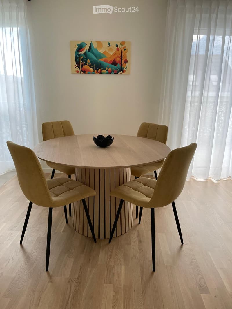 A modern dining area with a round wooden table surrounded by four beige upholstered chairs, a decorative bowl on the table, and a colorful abstract painting on the wall.