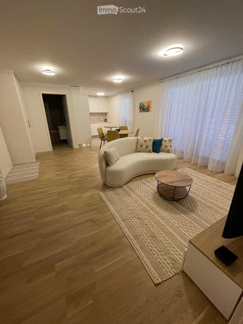 Modern living room with a curved white sofa, coffee table, dining area with chairs, large windows with curtains, and artwork on the wall.