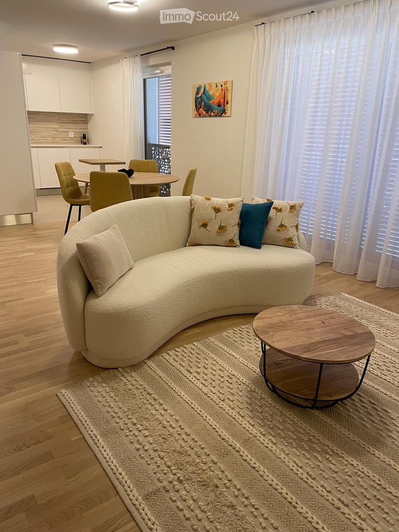 Modern living room with a curved sofa, coffee table, dining area, and large windows with white curtains.