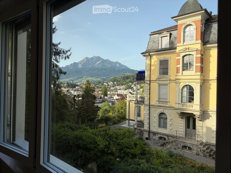 View from a window showing a large, yellow and white building with multiple stories and a dome-shaped roof, surrounded by greenery and trees, with a scenic mountain view in the background.